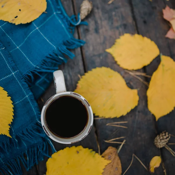 En una vieja mesa de madera en el parque de otoño hay una taza de té y café. Una bufanda a cuadros azul cálido se dispersa con hojas amarillas y conos de pino. Vista superior, borrosa. Otoño cálido estado de ánimo oscuro, espacio de copia — Foto de Stock