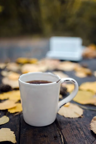 En una vieja mesa de madera en el parque de otoño hay una taza con té y café, hojas amarillas dispersas y conos de pino. Vista superior, borrosa. Otoño cálido humor oscuro, enfoque suave . — Foto de Stock