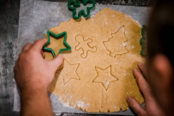 Male hands cut out gingerbread cookie in the form of a snowflake, Christmas tree, a man from raw dough on parchment baking paper on a dark background. View from above. save space — Stock Photo, Image