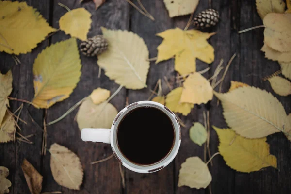 En una vieja mesa de madera en el parque de otoño hay una taza con té y café, hojas amarillas dispersas y conos de pino. Vista superior, borrosa. Otoño cálido humor oscuro, enfoque suave . — Foto de Stock