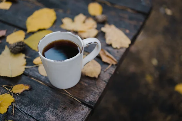 En una vieja mesa de madera en el parque de otoño hay una taza con té y café, hojas amarillas dispersas y conos de pino. Vista superior, borrosa. Otoño cálido humor oscuro, enfoque suave . — Foto de Stock