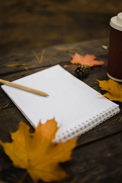 Белый пустой блокнот и ручка на темном фоне с шарфом, клеткой и чашкой, осенью желтые листья и шишки вокруг. Вид сверху. Плоская лежала, размытая. Копирование пространства Стоковое Изображение