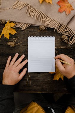 Ahşap masada, defter, kalem ve sonbahar yapraklarıyla birlikte bir fincan kahve ve eşarpla insan eli. Ahşap masada bir fincan kahve ve atkıyla insan eli.