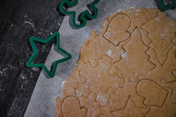 Corten la galleta de jengibre en forma del árbol de Navidad, la estrella, el hombrecito, los corazones de la masa cruda sobre el papel de hornear de pergamino sobre el fondo oscuro. Vista superior. ahorrar espacio — Foto de Stock