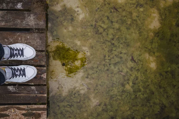 Top view of female legs in white sneakers and jeans on the background of an old dirty autumn lake-river and a wooden platform. Copy space — Stock Photo, Image