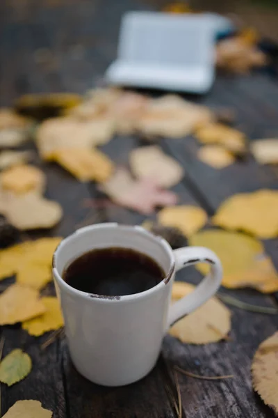En una vieja mesa de madera en el parque de otoño hay una taza con té y café, hojas amarillas dispersas y conos de pino. Vista superior, borrosa. Otoño cálido humor oscuro, enfoque suave . — Foto de Stock