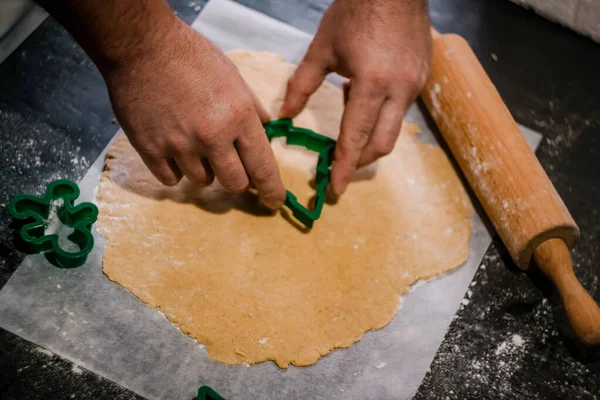 Male hands cut out gingerbread cookie in the form of a snowflake, Christmas tree, a man from raw dough on parchment baking paper on a dark background. View from above. save space — Stock Photo, Image