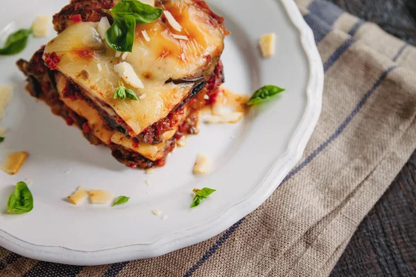 Tradicional PARMIGIANA di Melanzane: pieczone bakłażan-Włochy, sycylijska kuzynka. Pieczone bakłażan z serem, pomidorami i przyprawami na białej płytce. Danie z bakłażan jest na drewnianym stole — Zdjęcie stockowe