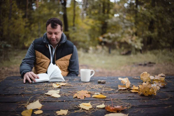 Genç bir adam sonbahar parkında eski ahşap bir masada oturur, çay içer ve kitap okur. Karanlık sonbahar atmosferi, yumuşak odaklanma. Bulanık olarak, boşluğu kopyala — Stok fotoğraf