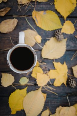 Sonbahar parkındaki eski ahşap bir masanın üzerinde çay, kahve, dağınık sarı yapraklar ve çam kozalakları olan bir fincan var. Üst manzara, bulanık. Sonbahar ılık karanlık ruh hali, yumuşak odaklanma.