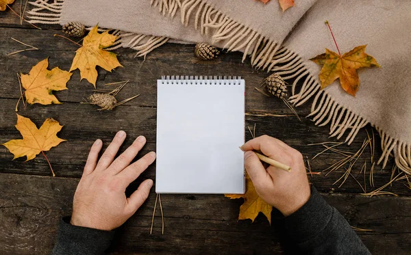 Menschenhände mit einer Tasse Kaffee und Schal am Holztisch mit Notizbuch und Stift und Herbstblättern. Menschenhände mit einer Tasse Kaffee und einem Schal am Holztisch — Stockfoto