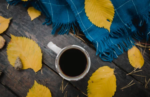 En una vieja mesa de madera en el parque de otoño hay una taza de té y café. Una bufanda a cuadros azul cálido se dispersa con hojas amarillas y conos de pino. Vista superior, borrosa. Otoño cálido estado de ánimo oscuro, espacio de copia — Foto de Stock