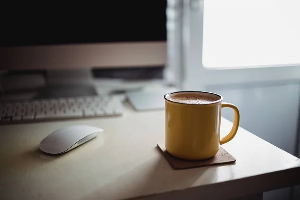 モニターとキーボードの横にある晴れた朝の光の中でオフィスの机の上にコーヒーの黄色のカップが立っています。 — ストック写真