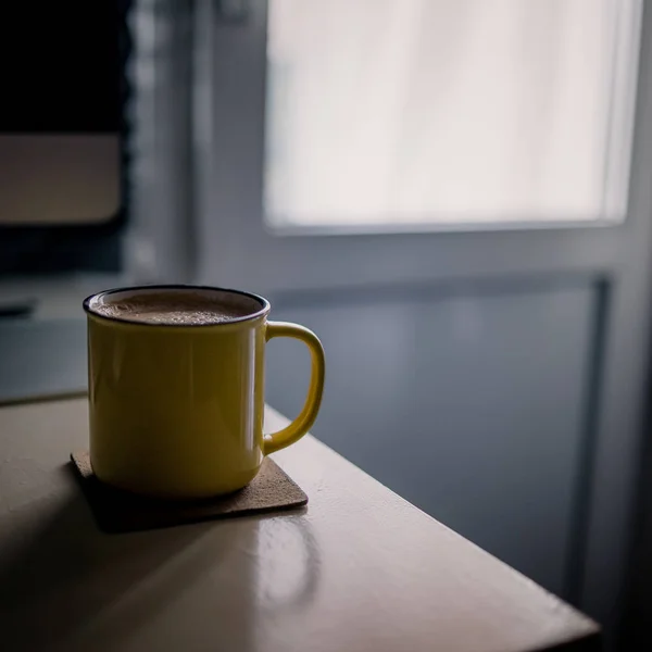 Eine gelbe Tasse Kaffee steht auf dem Schreibtisch im sonnigen Morgen- und Abendlicht neben Monitor und Tastatur — Stockfoto