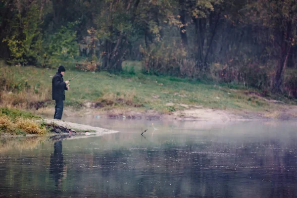 Visser in vervaging aan de oever van een meer met vishengel op een heldere herfstdag, reflectie in het water — Stockfoto
