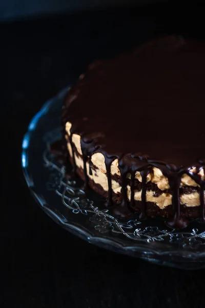 Chocolate round cake layers with butter-cream-nut cream with chocolate drips on a glass stand on a dark wooden background. Minimalism, Copy space. Selective focus, close-up.