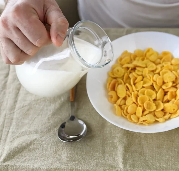 A levágott ember tejet önt a kukoricapelyhekbe egy fehér tányéron, egy sima, durva abroszon. Kilátás fentről. Szelektív fókusz. Koncepció, egyszerű gyors amerikai egészséges reggeli — Stock Fotó