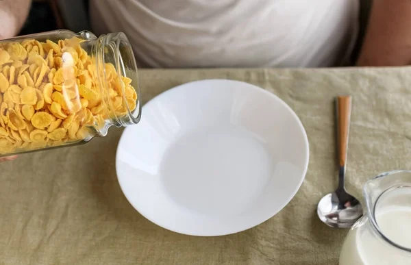 한 남자가 걸쭉하고 거친 식탁보 위에 하얀 접시에 콘플레이크를 뿌리고 있다. 클로즈업. 위에서 본 모습. 선택적 집중입니다. 콘셉트, 간단 한 미국 건강 아침 식사 — 스톡 사진