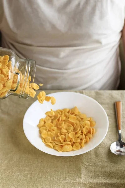 Un hombre recortado espolvorea copos de maíz en un plato blanco sobre un mantel liso y áspero. Primer plano. Vista superior. Enfoque selectivo. Concepto, desayuno saludable americano rápido simple — Foto de Stock