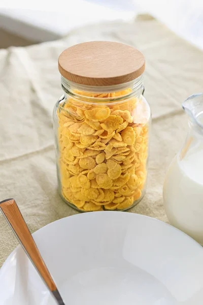 Copos de maíz en un frasco, junto a una jarra transparente de leche, un plato vacío con una cuchara sobre un mantel liso y áspero. Vista desde arriba . — Foto de Stock