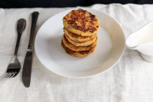 En la mesa con un mantel brillante de lino hay un barco de salsa blanca con salsa de crema agria, tortitas de queso cuajada en un plato, un tenedor, un cuchillo en un mantel liso blanco. Concepto, simple saludable — Foto de Stock