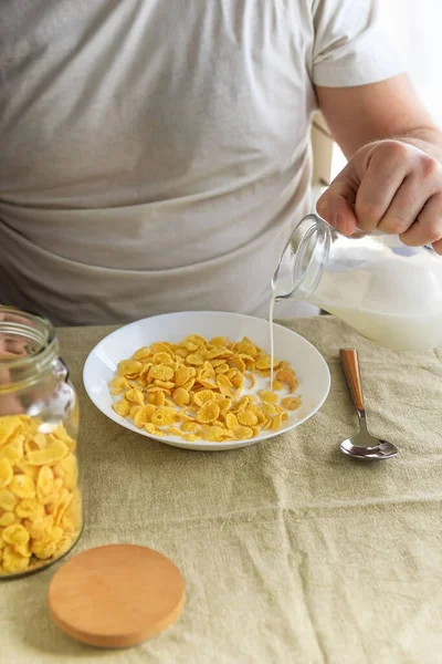 Кукурузник наливает молоко в кукурузные хлопья в белой тарелке на простой грубой скатерти. Вид сверху. Выборочный фокус. Концепция, простой быстрый американский здоровый завтрак — стоковое фото
