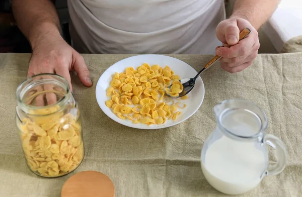절뚝거리는 남자가 곱고 거친 식탁보 위에 하얀 접시에 우유를 담은 콘플레이크 속에 수저를 들고 있다. 위에서 본 모습. 콘셉트, 간단 한 미국 건강 아침 식사 — 스톡 사진