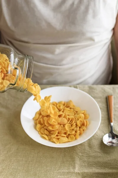 Un hombre recortado espolvorea copos de maíz en un plato blanco sobre un mantel liso y áspero. Primer plano. Vista superior. Enfoque selectivo. Concepto, desayuno saludable americano rápido simple — Foto de Stock