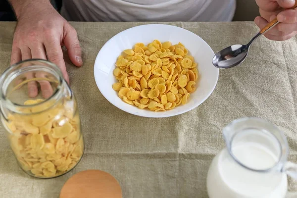 Egy levágott ember kanalat tart kukoricapelyhekben tejjel egy fehér tányéron egy sima, durva terítőn. Kilátás fentről. Koncepció, egyszerű gyors amerikai egészséges reggeli — Stock Fotó