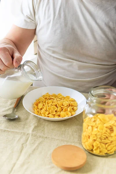 El hombre cortado vierte leche en copos de maíz en un plato blanco sobre un mantel liso y áspero. Vista desde arriba. Enfoque selectivo. Concepto, desayuno saludable americano rápido simple — Foto de Stock