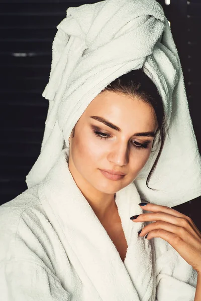 Sexy junges Mädchen mit dunklen Haaren, großen Augen und dunklen Augenbrauen im weißen Bademantel mit Handtuch auf dem Kopf. — Stockfoto