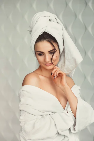 Sexy junges Mädchen mit dunklen Haaren, großen Augen und dunklen Augenbrauen im weißen Bademantel mit Handtuch auf dem Kopf. — Stockfoto