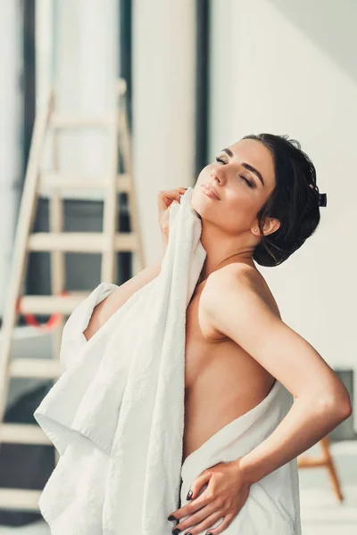 Sexy jonge brunette vrouw staande voor venster en het bewonderen van de zonsopgang dekking van haar naakte lichaam met handdoek na bad. — Stockfoto