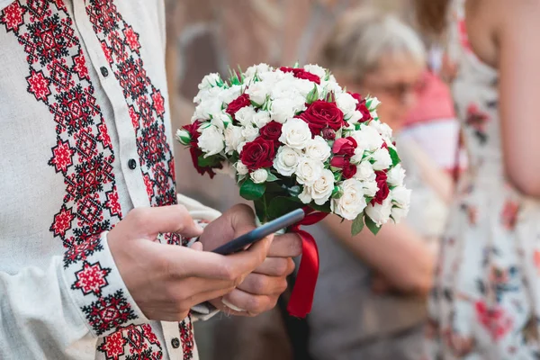 Narzeczony trzymamy w ręku telefon komórkowy i Bukiet ślubny z czerwonych róż i beż. — Zdjęcie stockowe