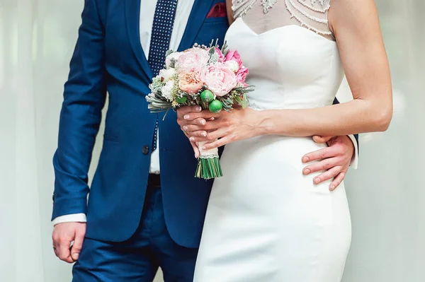 De mooie bruid houdt een bruiloft boeket met roze rozen en pioenrozen. Bruidegom omhelzing vrouw door de taille. — Stockfoto