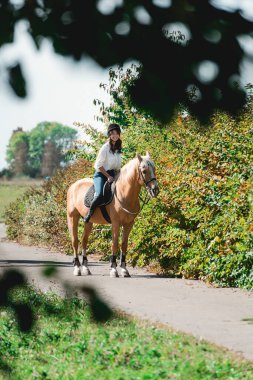 Beyaz gömlek ve miğfer giymiş at üstünde esmer bir kadının portresi. Ata binmeyi öğreniyor. Parkta binicilik dersleri.