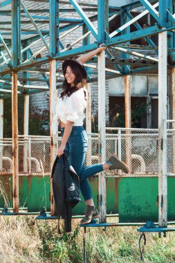 Kırsal metal yapısında poz veren güzel seksi kız. Beyaz gömlekli, siyah şapkalı ve kot pantolonlu bir kadın. Omzunda siyah ceket var.