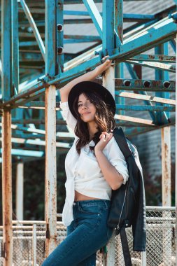 Kırsal metal yapısında poz veren güzel seksi kız. Beyaz gömlekli, siyah şapkalı ve kot pantolonlu bir kadın. Omzunda siyah ceket var.