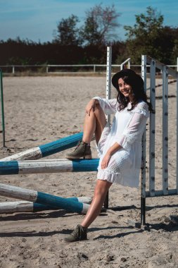 Güzel, seksi bir kız at epodrom 'unda yatay olarak poz veriyor. Beyaz elbiseli, siyah şapkalı ve siyah çizmeli bir kadın. Yaz zamanı.