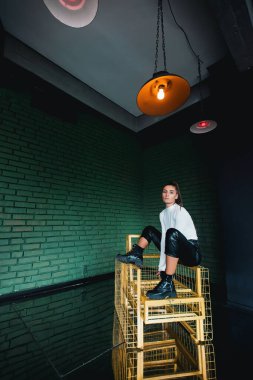 Beyaz kazaklı, siyah deri pantolonlu, yeşil tuğlalı arka planda güneş ışığında poz veren şık esmer kız..