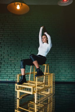 Beyaz kazaklı, siyah deri pantolonlu, yeşil tuğlalı arka planda güneş ışığında poz veren şık esmer kız..