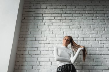 Beyaz kazaklı ve siyah deri pantolonlu şık esmer kız beyaz tuğlalı arka planda güneş ışığında poz veriyor..