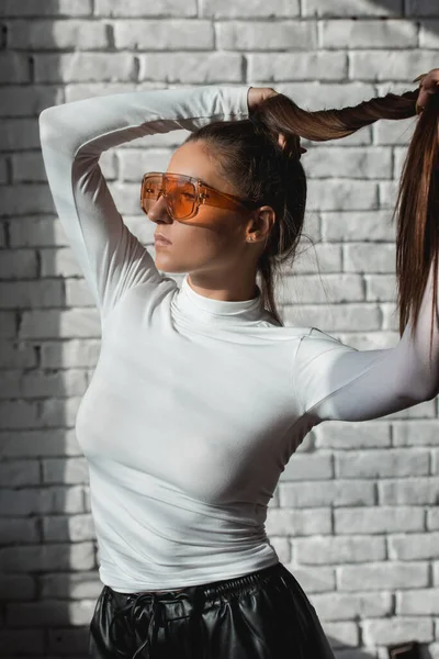 Fashionabla brunett flicka i en vit tröja, orange glasögon och svart läder byxor poserar på en vit tegelsten bakgrund. — Stockfoto