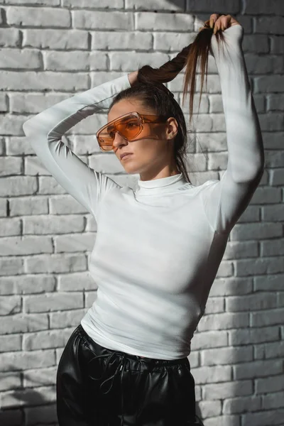 Menina morena na moda em um suéter branco, óculos de laranja e calças de couro preto posando em um fundo de tijolos brancos . — Fotografia de Stock