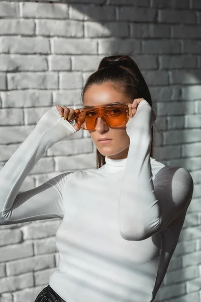 Menina morena na moda em um suéter branco, óculos de laranja e calças de couro preto posando em um fundo de tijolos brancos . — Fotografia de Stock