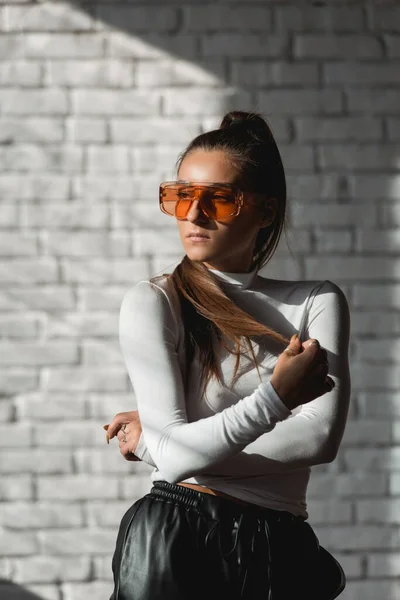 Ragazza bruna alla moda in un maglione bianco, occhiali arancioni e pantaloni di pelle nera in posa su uno sfondo di mattoni bianchi . — Foto Stock