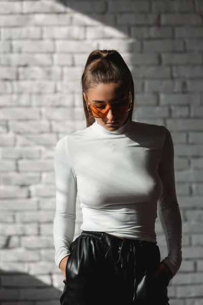 Ragazza bruna alla moda in un maglione bianco, occhiali arancioni e pantaloni di pelle nera in posa su uno sfondo di mattoni bianchi . — Foto Stock