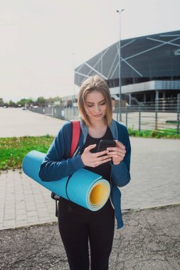 Tonlu genç yoga eğitmeni ayakta durup akıllı telefondan postaları kontrol ediyor, antrenmana ara veriyor, spor giyim sektöründe milenyum bayan sporcusu. Açık havada eğitim için cep telefonu kullanıyor..
