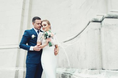 Beyaz gelinlikli sarı saçlı gelin ve beyaz antik bir binanın arka planında takım elbiseli damat. Gelin düğün buketi tutuyor.