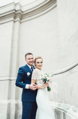 Beyaz gelinlikli sarı saçlı gelin ve beyaz antik bir binanın arka planında takım elbiseli damat. Gelin düğün buketi tutuyor.